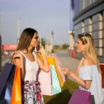 Two fashionable women carry colorful shopping bags in a sunny urban environment.